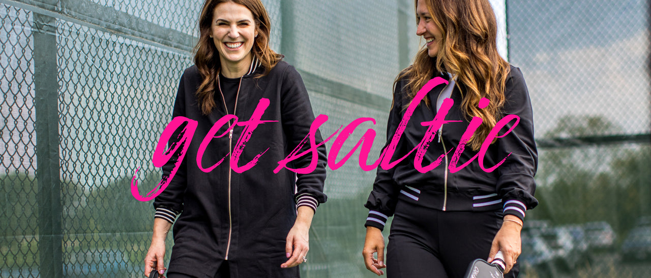  Two women pickleball players in matching black Saltie Rose outfits with pink text 'get salty' standing in front of a chain-link fence.